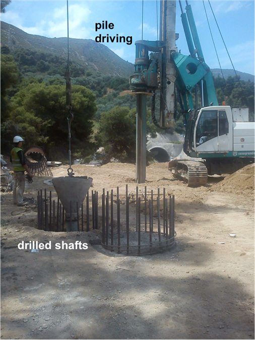 Photograph of a pile driving machine installing an H-shaped steel pile. Next to the driven pile there are two drilled shafts, with the reinforcement cage extruding. Concrete is being poured into one of the drilled shafts through a tremie.